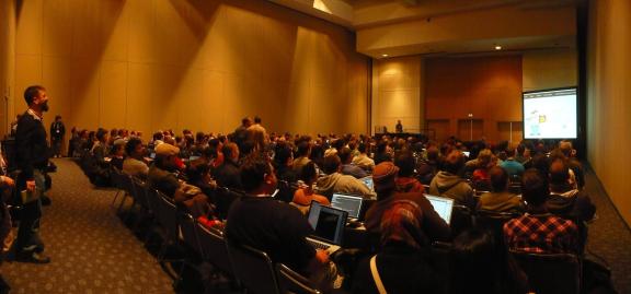 A wide view of a packed conference session room with an audience facing a projector screen.