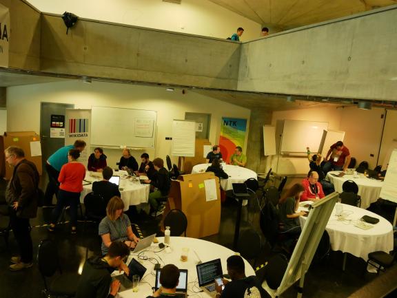 People working at round tables with laptops in a large, bright hacking space.