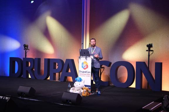 A speaker on the main stage at DrupalCon Rotterdam 2026, in front of large DrupalCon letters lit in blue and gold.