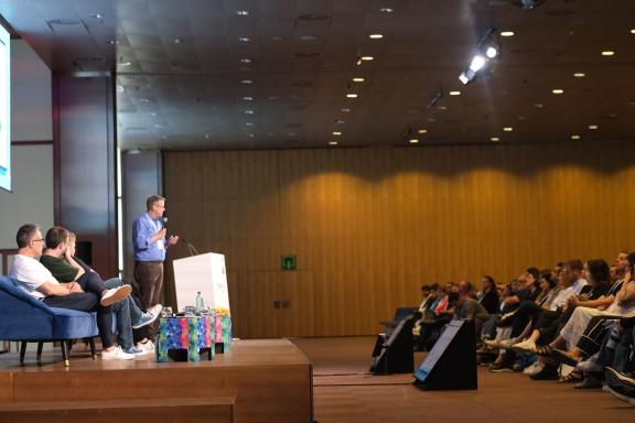 A speaker standing beside a seated panel on stage, facing an audience at DrupalCon Barcelona 2024.