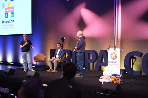 Three people on the main stage at DrupalCon Rotterdam 2026, one speaking into a microphone, with the conference logo on a screen behind them.