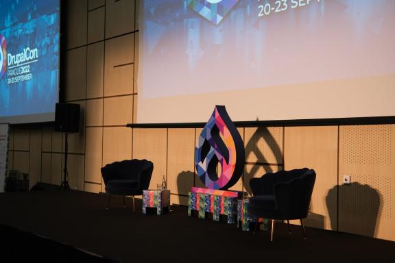 An empty conference stage with two armchairs and a large Drupal drop sculpture, lit warmly before a session.