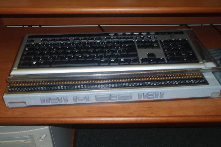 A braille display sitting in front of a computer keyboard on a wooden desk.