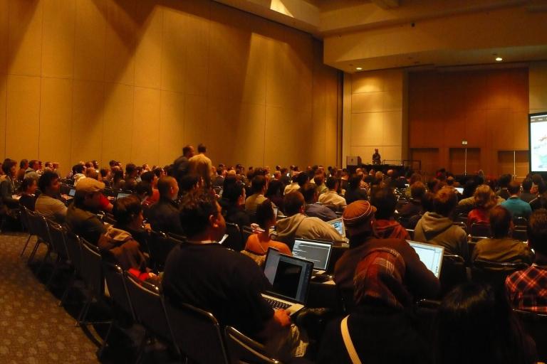A wide view of a packed conference session room with an audience facing a projector screen.