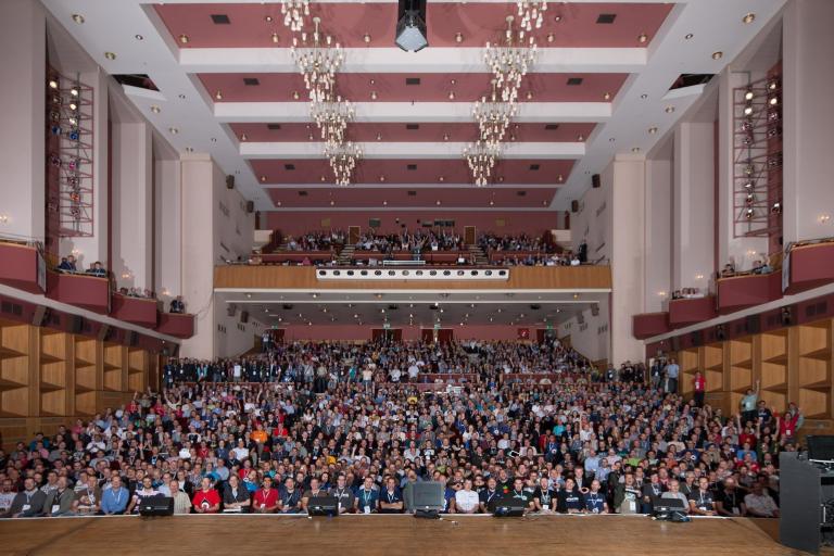 A large group photo of conference attendees filling an ornate concert hall.