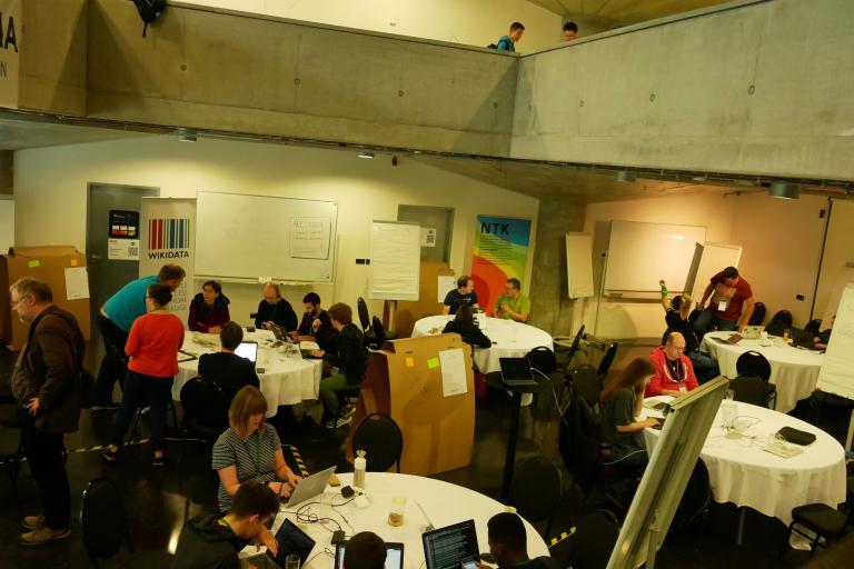 People working at round tables with laptops in a large, bright hacking space.