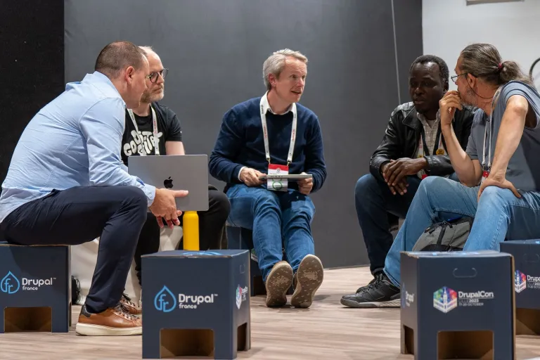 Five people sitting on cardboard stools, deep in conversation around a laptop at DrupalCon Lille 2023.