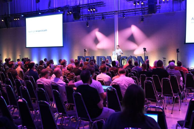 A large audience facing the main stage at DrupalCon Rotterdam 2026, where a speaker stands in front of large letters spelling DrupalCon.