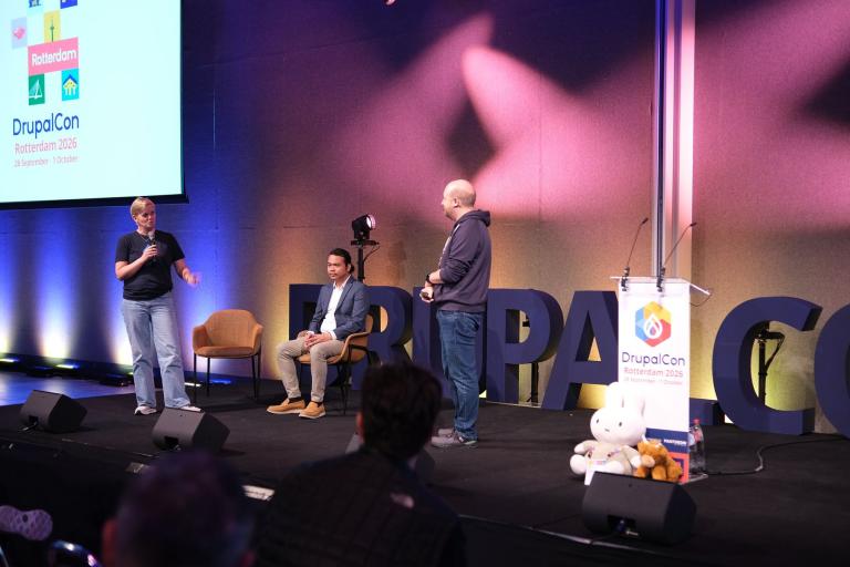 Three people on the main stage at DrupalCon Rotterdam 2026, one speaking into a microphone, with the conference logo on a screen behind them.