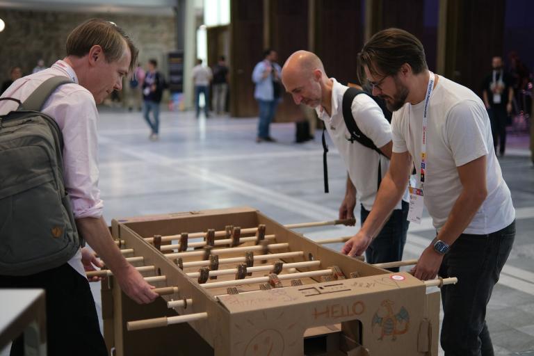Three attendees playing on a cardboard table football set in the hall at DrupalCon Rotterdam 2026.