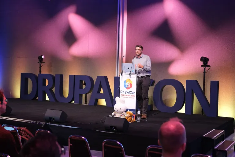 A speaker at a lectern on a stage, in front of large letters spelling DrupalCon.