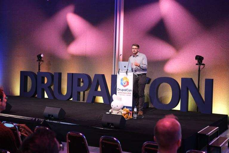 A speaker at a lectern on a stage, in front of large letters spelling DrupalCon.